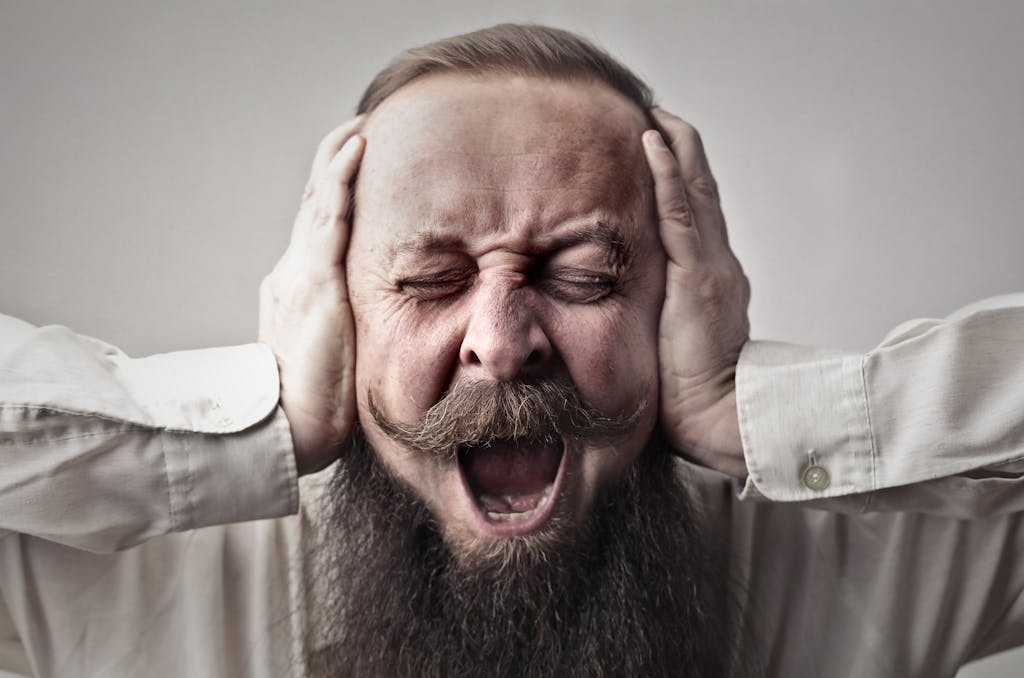 Portrait of a man with facial hair, covering his ears and screaming, expressing stress or frustration.